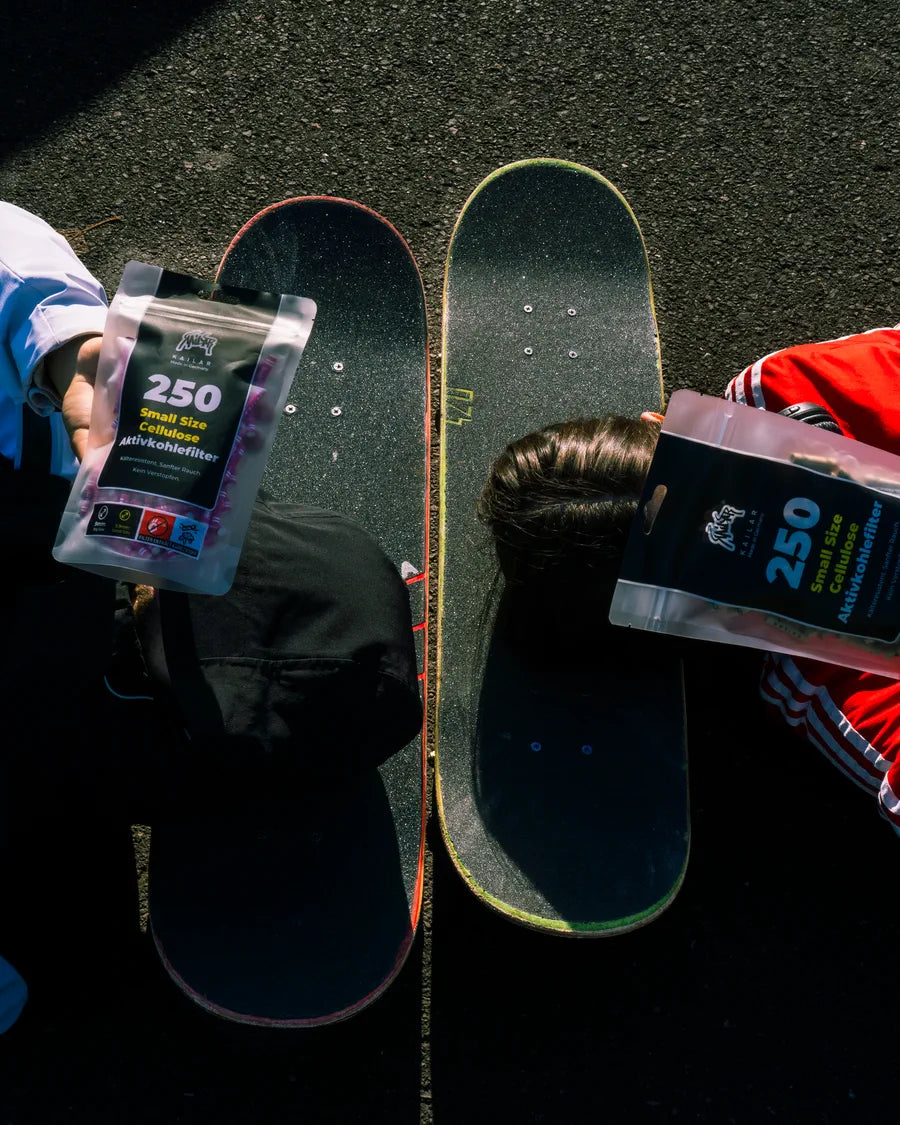 Deux skateboards sur l’asphalte, à côté deux personnes tiennent chacune un paquet de 250 filtres Kailar
